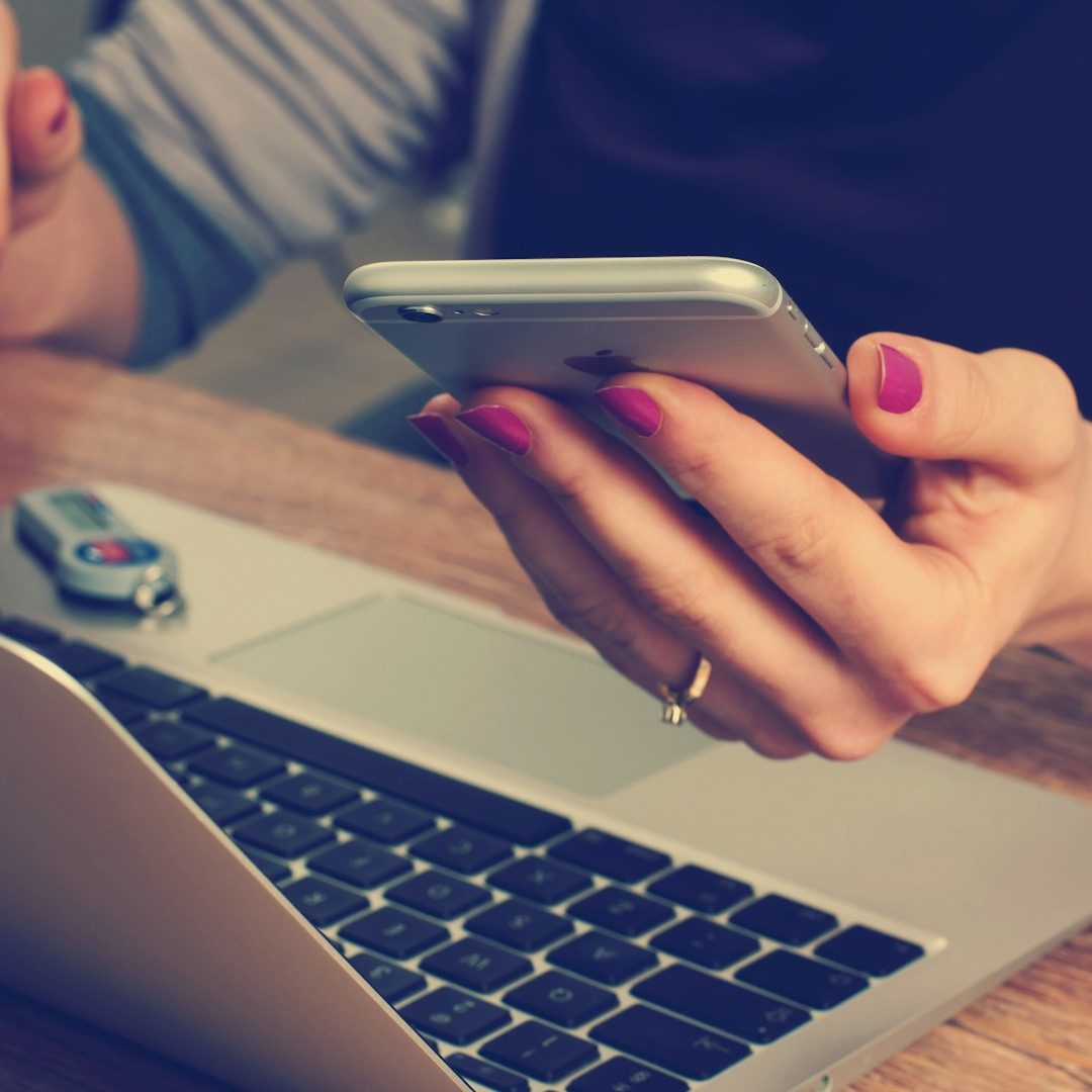 Eine Hand hält ein Smartphone, während die andere auf einem Laptop tippt.